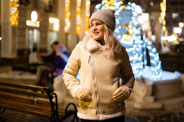 Straßenporträt der lächelnden schönen jungen Frau auf dem festlichen Weihnachtsmarkt. Dame, die klassische, stilvolle Winterstrickkleidung trägt.