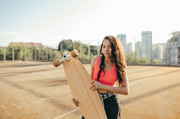Straßenporträt des attraktiven lockigen Mädchens mit Skateboard