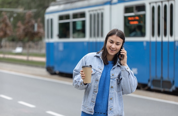 Straßenporträt einer fröhlichen jungen Frau, die mit Kaffee zu ihrer Hand telefoniert.