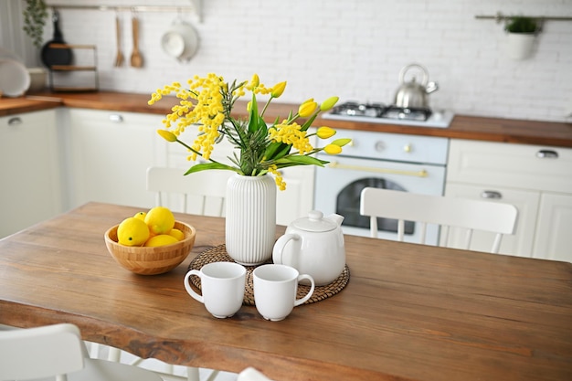 Strauß gelber Tulpen und Mimosen in einer Vase auf einem Holztisch vor dem Hintergrund eines weißen Mo...