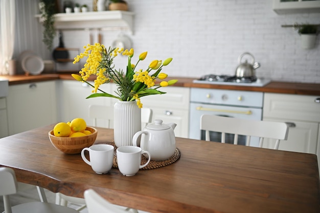 Strauß gelber Tulpen und Mimosen in einer Vase auf einem Holztisch vor dem Hintergrund eines weißen Mo...