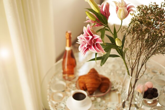 Strauß rosa Lilien. Tasse Kaffee, Croissants und eine Flasche Wein auf unscharfem Hintergrund. Festliches Frühstück. Licht positives Foto