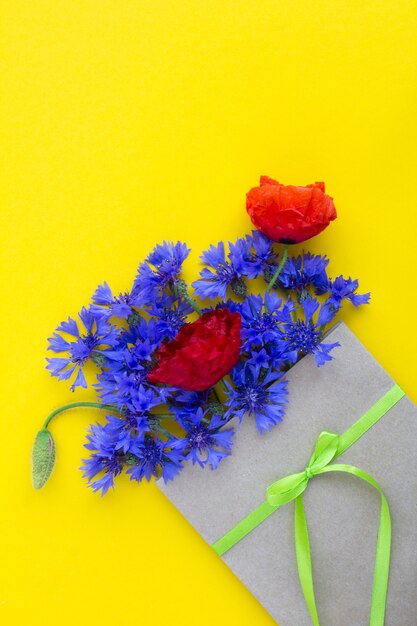 Strauß roter Mohnblumen und blauer Kornblumen in einem Umschlag gebunden mit einem grünen Band auf der gelben Oberfläche. Draufsicht. Kopierraum. Frühlings- oder Sommerblumenkonzept.