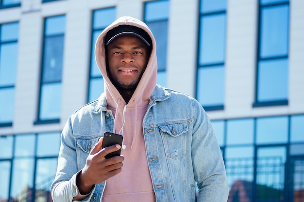 Street-Fashion-Konzept - stilvoller, gutaussehender afroamerikanischer Mann in rosa Hemdkapuze und Jeansjacke und schwarzer Mütze vor den Fenstern des Geschäftszentrums