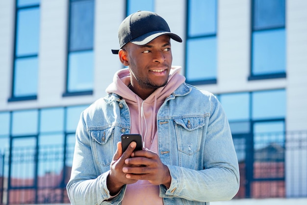 Street-Fashion-Konzept - stilvoller, gutaussehender afroamerikanischer Mann in rosa Hemdkapuze und Jeansjacke und schwarzer Mütze vor den Fenstern des Geschäftszentrums