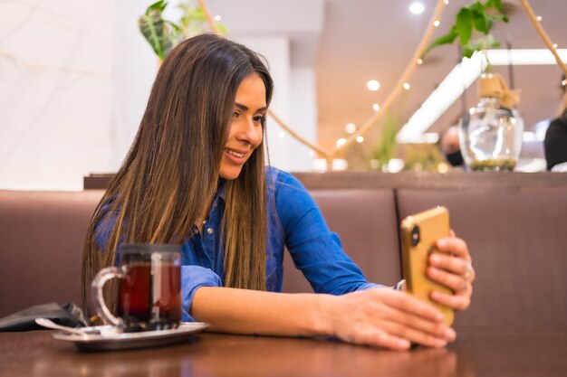 Foto street style en la ciudad, una chica morena de raza blanca con una camisa de mezclilla tomando el té en una cafetería. hacer un directo en las redes sociales con el teléfono