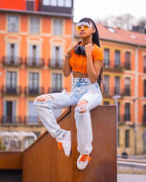 Street Style eines jungen Trap-Tänzers mit Zöpfen. Ein schönes schwarzes Mädchen der afrikanischen ethnischen Gruppe mit T-Shirt, orange Sonnenbrille und Cowboyhosensitzen. Mit orangefarbenem Häuserhintergrund