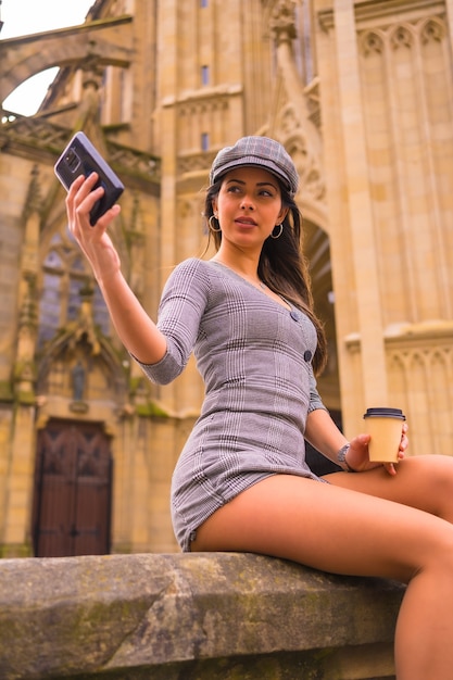 Street Style in der Stadt, brünettes kaukasisches Mädchen mit grauem Kleid und Baskenmütze, neben einer Kathedrale in der Stadt, die ein Selfie macht, Lebensstil eines Modells in der Stadt, vertikales Foto