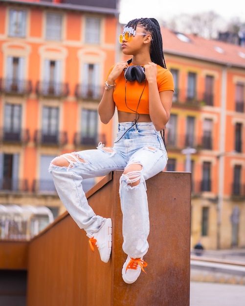 Street Style de una joven bailarina de trap con trenzas. Una hermosa chica negra de etnia africana con camiseta, gafas de sol naranjas y pantalones de vaquero sentado. Con fondo de casas naranja