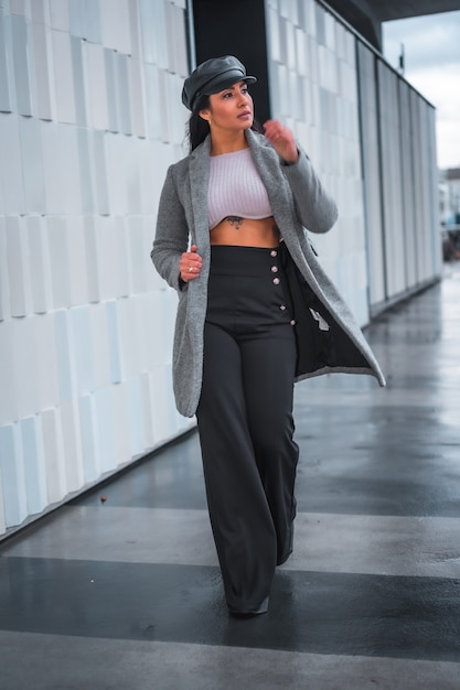 Street style na cidade de uma menina morena latina com um chapéu de couro e uma capa de chuva cinza andando pela cidade