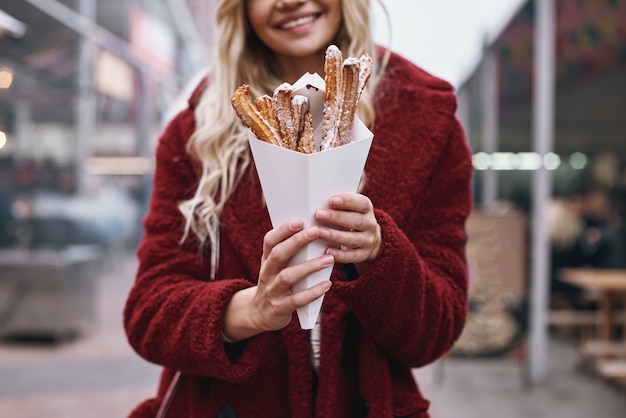 Streetfood-Kultur. Junge blonde Frau in roter Mütze und Öko-Pelzmantel, die Churros auf der Straßenmesse isst.