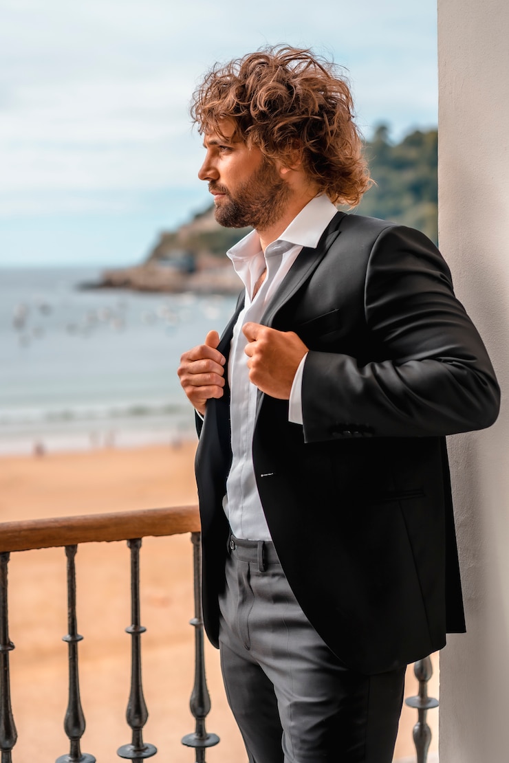 Foto streetstyle mit einem jungen brünetten kaukasischen mann in einem schwarzen anzug und einem weißen hemd in einem luxuriösen hotel, mode-editorial. modell sitzt am fenster