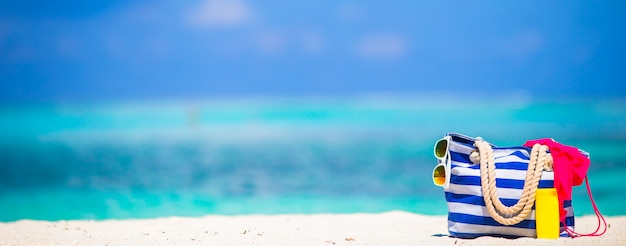 Streifentasche, blaues Tuch, Sonnenbrille, Sonnenschutzflasche und Badeanzug auf weißem Strand