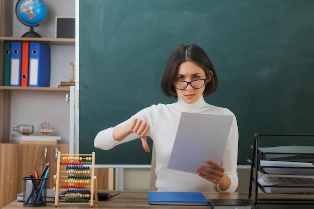 Strenge Daumen nach unten Junge Lehrerin mit Brille und Papier am Schreibtisch sitzend mit Schulwerkzeugen im Klassenzimmer