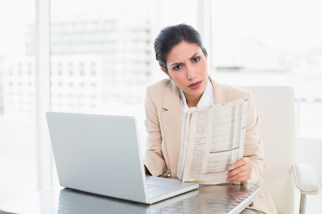 Strenge Geschäftsfrau, die Zeitung beim Arbeiten an dem Laptop betrachtet Kamera hält