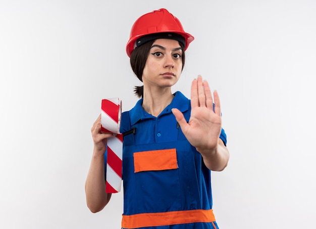 Strenge junge Baumeisterin in Uniform mit Klebeband, die Stoppgeste zeigt