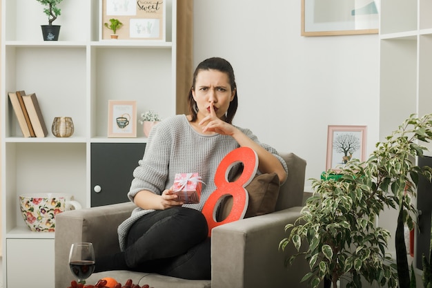 Strenge zeigende Stille Geste schönes Mädchen am glücklichen Frauentag mit Geschenk auf Sessel im Wohnzimmer sitzend