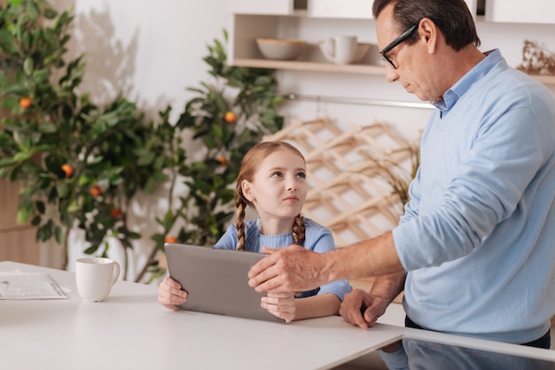 Strenger, selbstbewusster Großvater, der zu Hause steht, während er der Enkelin und dem kleinen Kind ein digitales Gerät wegnimmt