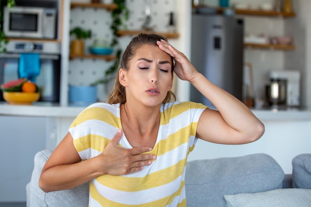 Stressige junge Frau, die Schmerzen fühlt und die Brust berührt, leidet zu Hause an Herzschmerzen, während sie einen Herzinfarkt hat
