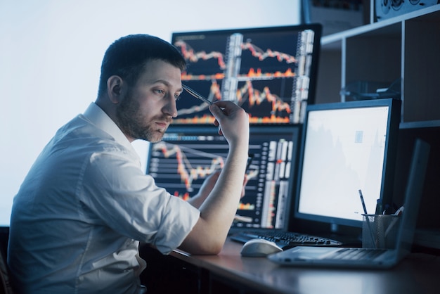 Stressiger Tag im Büro. Händchenhalten des jungen Geschäftsmannes auf seinem Gesicht beim Sitzen am Schreibtisch im kreativen Büro. Börse, die Devisen-Finanzgraphik handelt