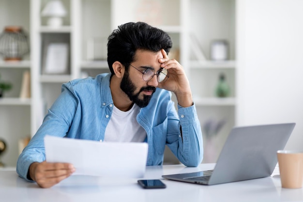 Stresster junger indischer Mann schaut auf den Laptop-Bildschirm und hält Dokumente in der Hand