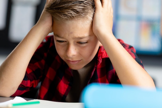 Stresster kaukasischer Schuljunge sitzt mit dem Kopf in den Händen am Schreibtisch und starrt im Unterricht auf das Buch