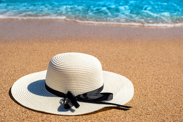 Strohhut der weißen Frau, der auf tropischem Sandstrand mit blauem lebendigem Ozeanwasser im Hintergrund am sonnigen Sommertag liegt. Urlaubs- und Reisezielkonzept.