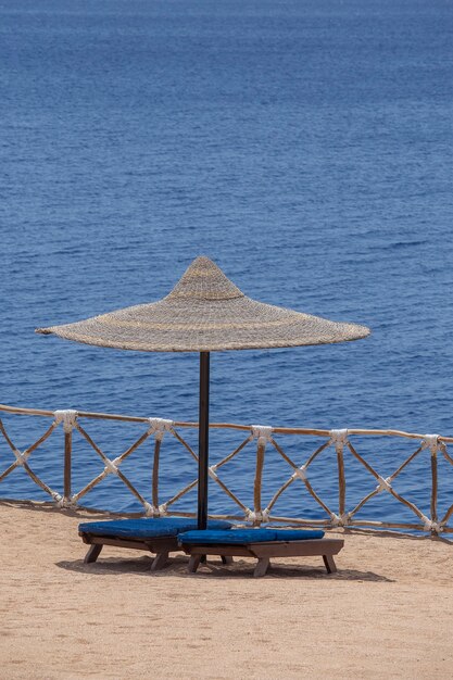 Strohschirme mit hölzernen Sonnenliegen neben dem roten Meerwasser am Sandstrand im Resort in Sharm El Sheikh, Ägypten