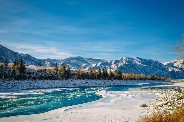 Strom des Flusses zwischen schneebedeckten Ufern gegen Berg mit Schnee und blauem Himmel an sonnigem frostigem Tag