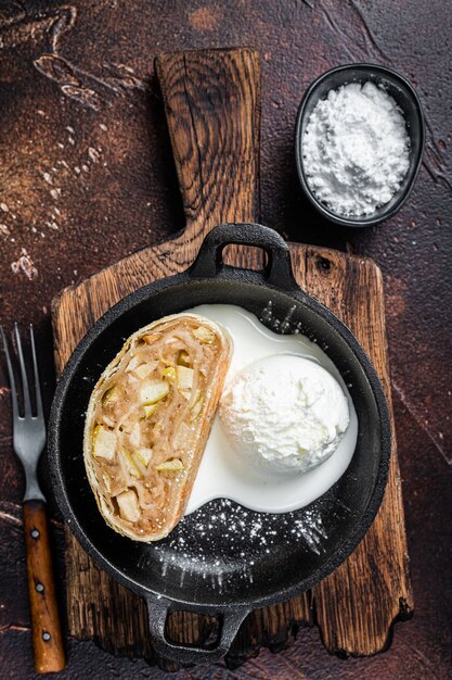 Strudel de apfelstrudel con canela, azúcar glass y helado de vainilla en una sartén. Fondo oscuro. Vista superior.