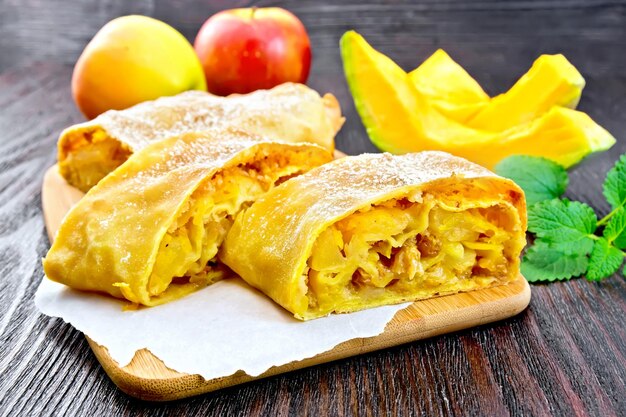 Strudel de calabaza y manzana con pasas sobre pergamino, menta, frutas y verduras sobre una tabla de madera oscura.