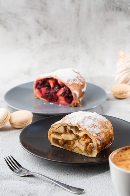 Strudel de maçã austríaco tradicional caseiro com maçãs frescas, nozes e açúcar de confeiteiro. menu para café. pedaço de bolo na chapa preta, copo branco sobre fundo branco de mármore. foto vertical.