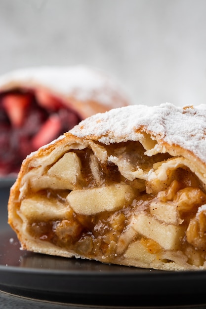 Strudel de maçã austríaco tradicional caseiro com maçãs frescas, nozes e açúcar de confeiteiro. Menu para café. Pedaço de bolo na chapa preta, copo branco sobre fundo branco de mármore. Foto vertical.