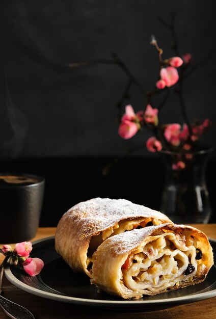 Strudel de manzana con nueces y pasas, vapor saliendo de una taza de espresso. En el fondo, una rama floreciente de un arbusto de primavera, primer plano, enfoque selectivo. Hornear para el fin de semana, vacaciones de primavera.