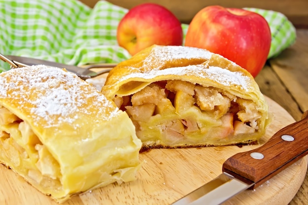 Strudel con manzanas, manzanas, un cuchillo, un colador, una servilleta en el fondo de tablas de madera