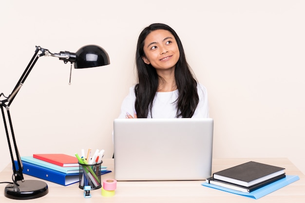 Student asiatisches Mädchen in einem Arbeitsplatz mit einem Laptop lokalisiert auf beigem Hintergrund, der beim Lächeln nach oben schaut