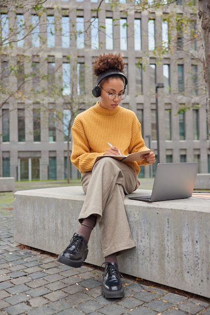 Student benutzt Laptop-Computer zum Lernen bereitet sich auf Vorlesungen an der Universität vor schreibt Notizen in Heft hört Webinar mit drahtlosen Kopfhörern posiert gegen städtische Umgebung