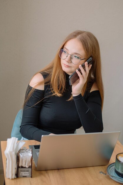 Student, der aus der Ferne arbeitet Eine junge Frau in einer schwarzen Bluse arbeitet an einem Laptop, telefoniert und trinkt Kaffee in einem Café Student, der aus der Ferne arbeitet