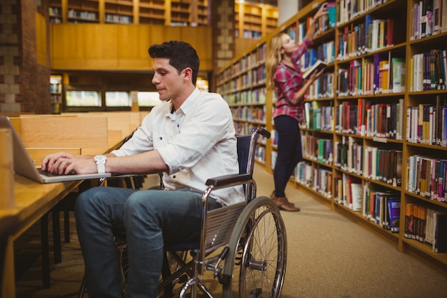 Student im Rollstuhl schreibend auf seinem Laptop während Frau, die Bücher in der Bibliothek sucht