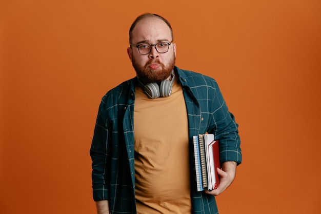 Student in lässiger Kleidung mit Brille und Kopfhörern, die Notizbücher in die Kamera blicken und mit traurigem Ausdruck über orangefarbenem Hintergrund stehen