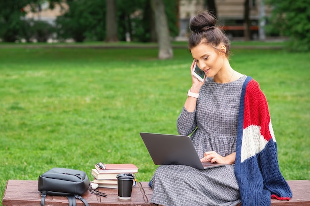 Student junge Frau spricht per Smartphone mit Laptop auf der Bank im Park