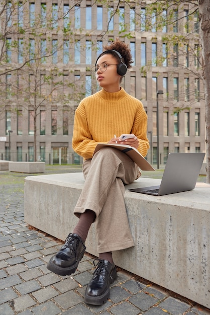 Student mit Afro-Haarstudien im Freien hört Bildungsvortrag über Kopfhörer auf schreibt Notizen im Notebook verwendet Laptop-Computer, um Projektposen gegen moderne Gebäude zu machen