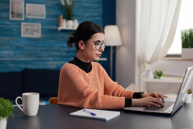 Student mit Brille, die auf dem Laptop sitzt, der am Schreibtisch zu Hause sitzt. College-Mädchen mit Notebook, das Fernbedienung mit Blick auf den Bildschirm studiert. Freiberufler, der von zu Hause aus E-Mails sendet. Programmierer, der im Internet surft.