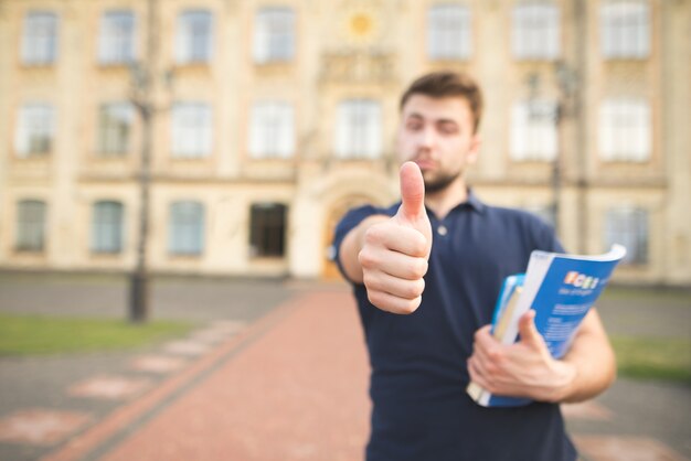 Student mit Büchern in den Händen steht auf dem Campus der Universität und zeigt einen Daumen hoch.