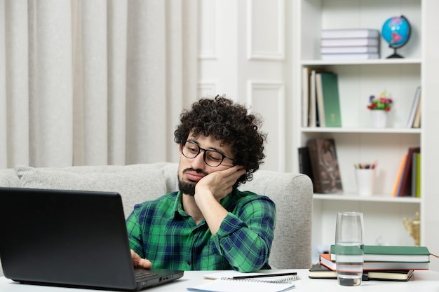 Student online süßer junger Kerl, der am Computer mit Brille im grünen Hemd studiert, müde studiert