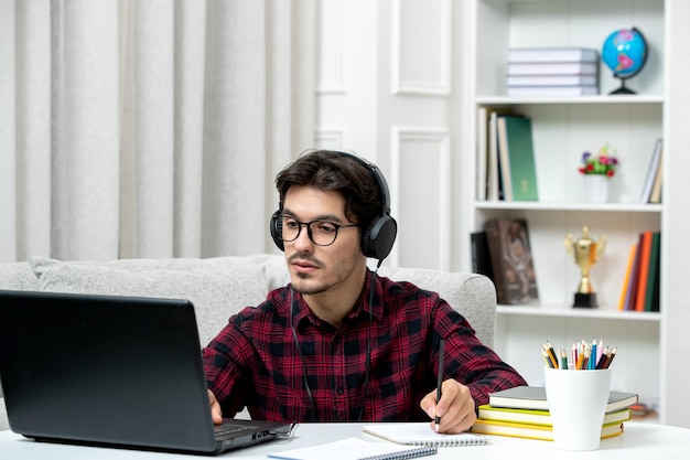 Student online süßer Typ in kariertem Hemd mit Brille, der am Computer studiert und Notizen schreibt