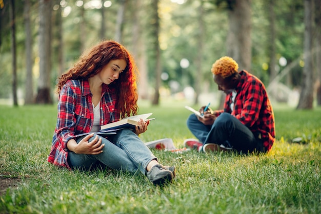 Studenten lesen Buch auf dem Rasen im Sommerpark. Männliche und weibliche Teenager, die draußen studieren und zu Mittag essen