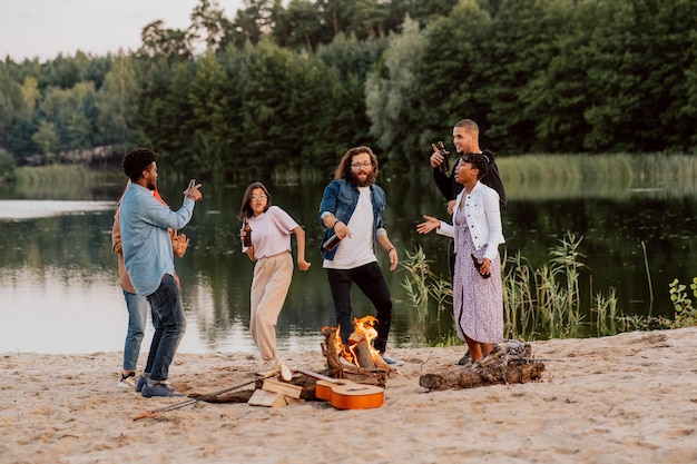 Studentenfreunde treffen sich am Strand am See, zünden ein Lagerfeuer an, braten Brotwürste, spielen Gitarre