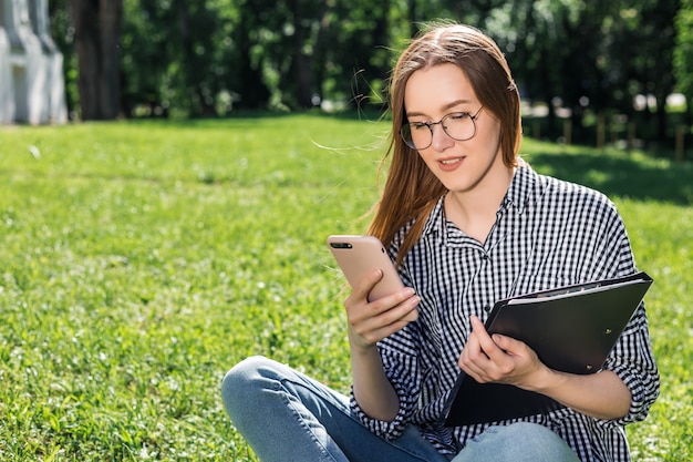 Studentenmädchen untersucht ihren Smartphone mit einem Ordner in ihren Händen, die auf dem Gras im Park sitzen