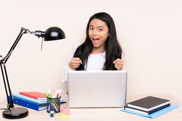 Studentin asiatisches Mädchen in einem Arbeitsplatz mit einem Laptop lokalisiert auf beige Wand, die einen Sieg in der Siegerposition feiert